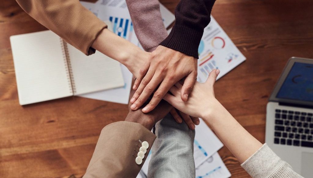 Image of six hands huddled in a circle over a conference room table for an article about Strategic Sourcing Solutions to Enhance Your Bottomline.